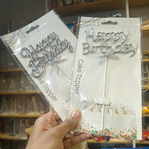 Two packaged silver 'Happy Birthday' cake toppers held in a hand, with shelves of similar items in the background.