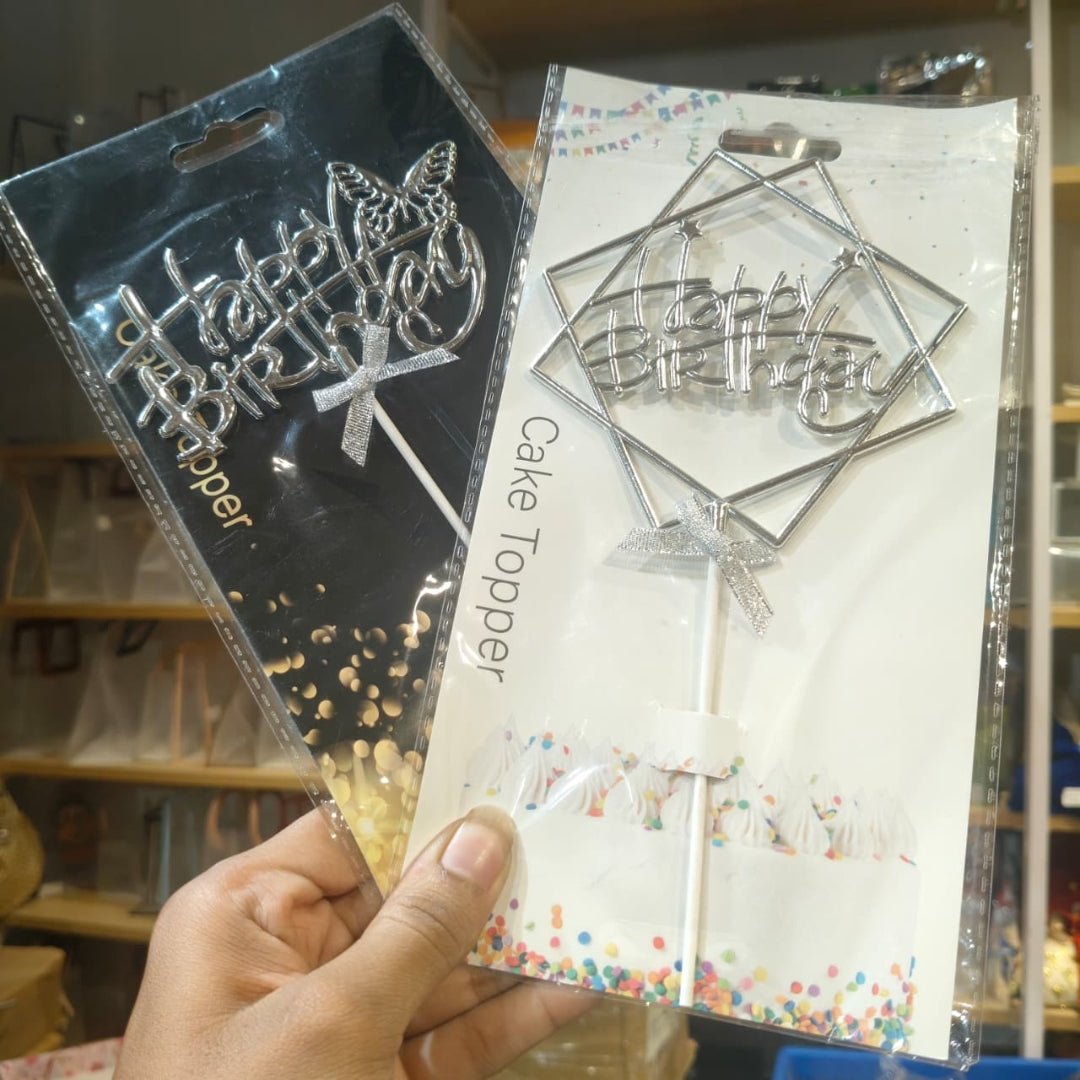 A hand holds two packaged silver cake toppers with the words "Happy Birthday" in decorative script and geometric frames, shown against a blurred store background.