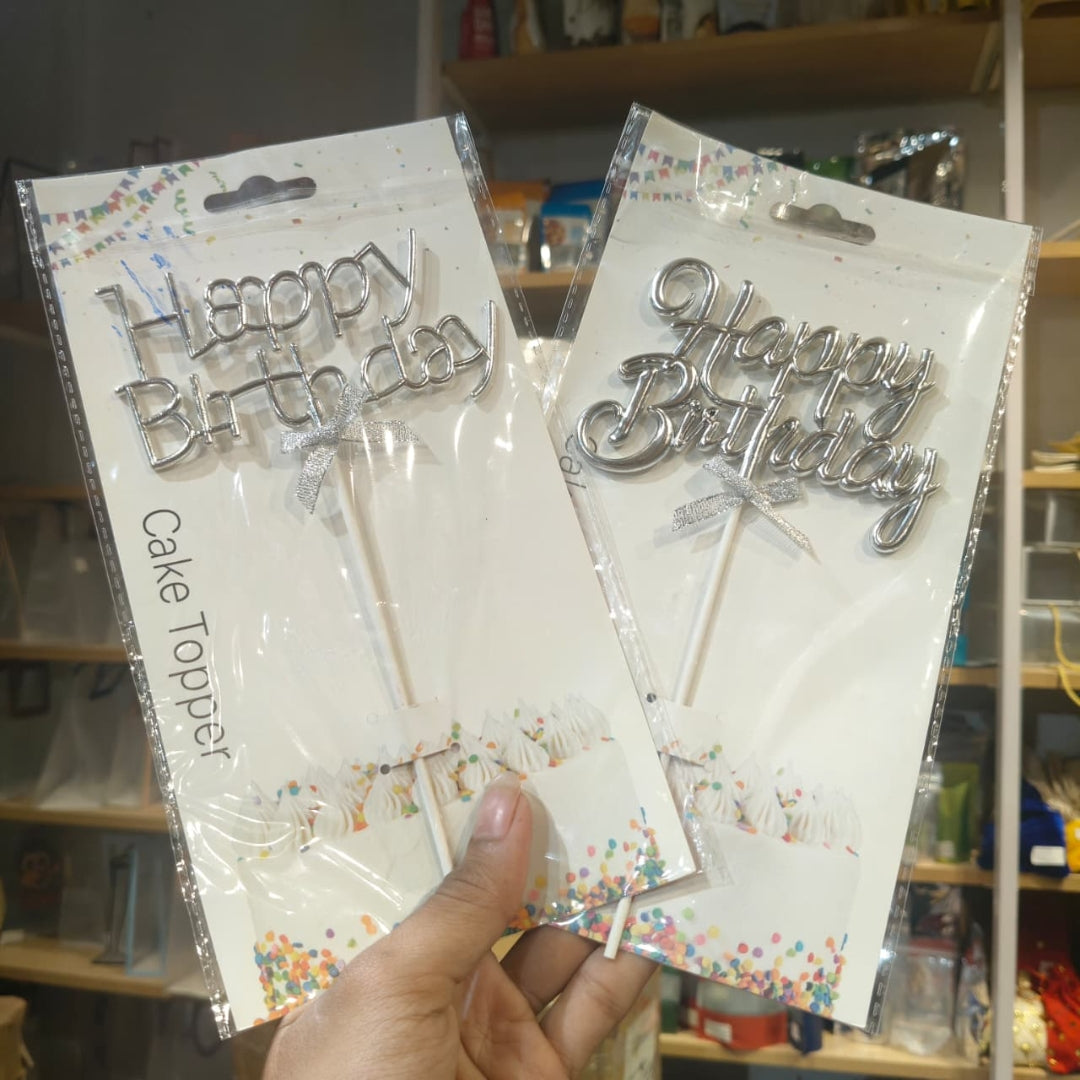Two packaged silver metal 'Happy Birthday' cake toppers held in a hand, with white backing cards and colorful confetti graphics, photographed in a store setting.