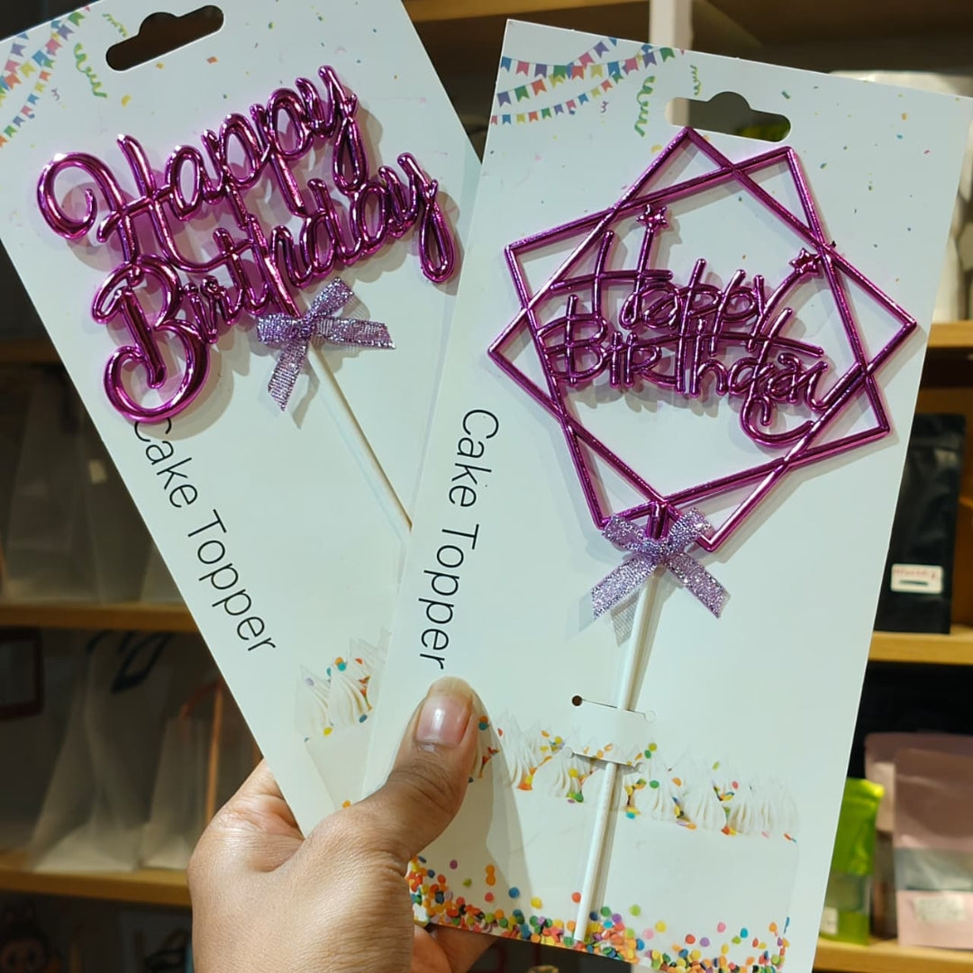 Two packaged 'Happy Birthday' cake toppers held in a hand. One is a shiny pink script letter cake topper on a white card, and the other is a metallic magenta geometric wire frame cake topper with the same script text on a white card.