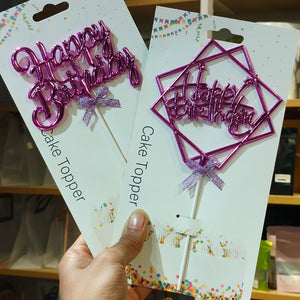 Two packaged 'Happy Birthday' cake toppers held in a hand. One is a shiny pink script letter cake topper on a white card, and the other is a metallic magenta geometric wire frame cake topper with the same script text on a white card.