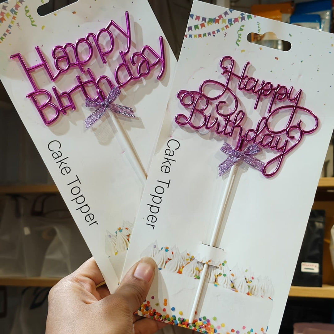 Two packaged pink glitter 'Happy Birthday' cake toppers held in a hand, with white retail cards showing the product and confetti illustrations.