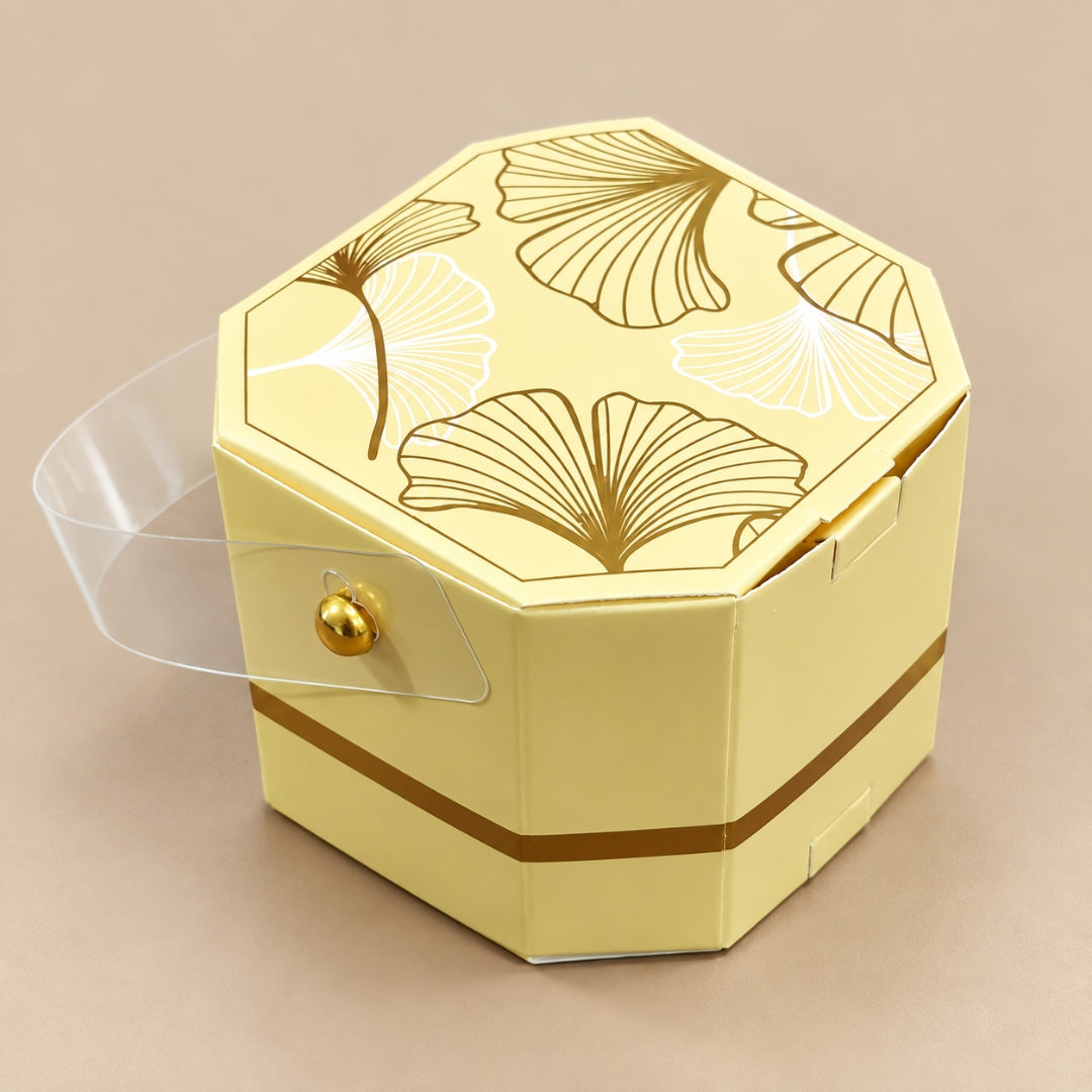 A pale yellow hexagonal gift box with a clear plastic hinged lid, decorated with gold and white ginkgo leaf patterns on the lid and a gold band around the lower side, set against a solid beige background.