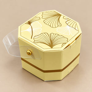 A pale yellow hexagonal gift box with a clear plastic hinged lid, decorated with gold and white ginkgo leaf patterns on the lid and a gold band around the lower side, set against a solid beige background.