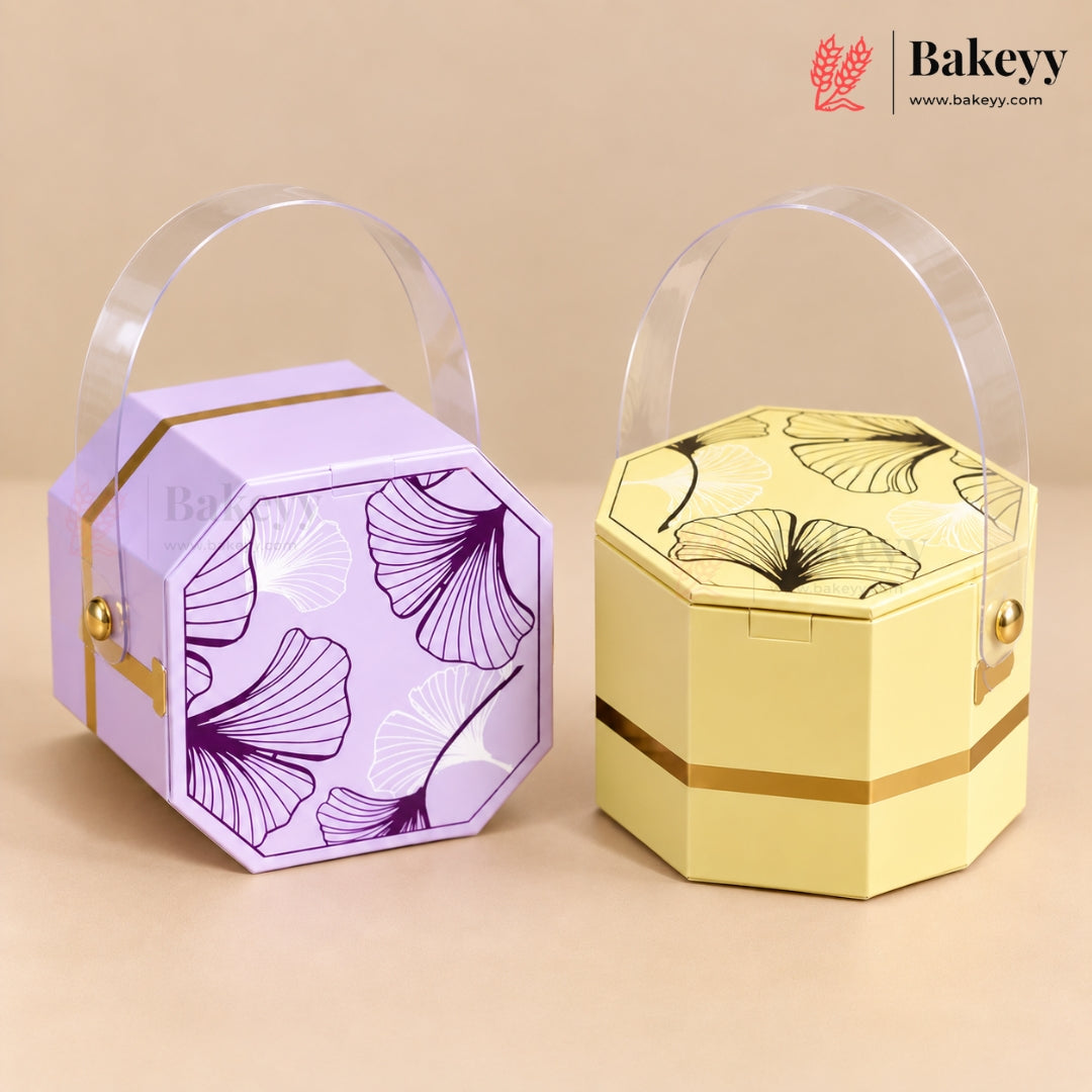 Two octagonal bakery gift boxes with clear plastic handles, shown on a beige background. The left box is lavender purple with purple ginkgo leaf patterns and gold trim.