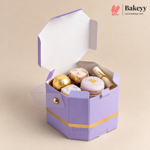A lavender and gold hexagonal cardboard gift box with a clear hinged lid, shown open and filled with assorted pastries including macarons and chocolate-covered spheres. The Bakeyy brand logo is visible in the top right corner.