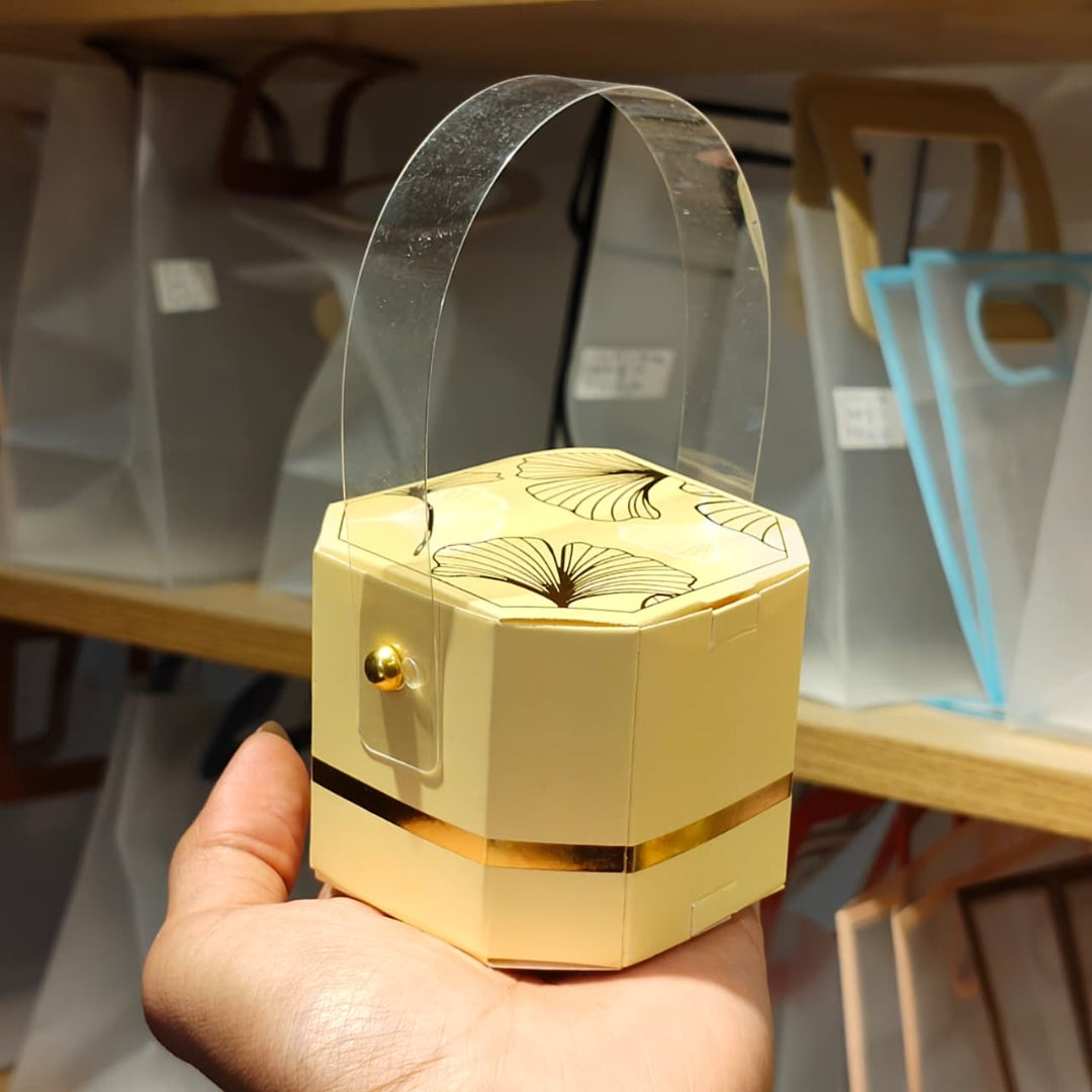 A hand holding a small hexagonal cream-colored gift box with a clear plastic handle and gold accents, with blurred clear plastic bags on wooden shelves in the background.