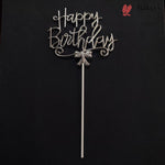 A silver metal 'Happy Birthday' cake topper with a bow detail and long stick, displayed against a solid black background. A Bakey bakery logo is visible in the top right corner.