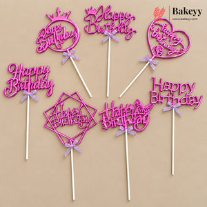 Six pink 'Happy Birthday' cake toppers with white sticks and small lavender ribbon bows, arranged on a beige background. The toppers include crown, heart, and geometric shapes.