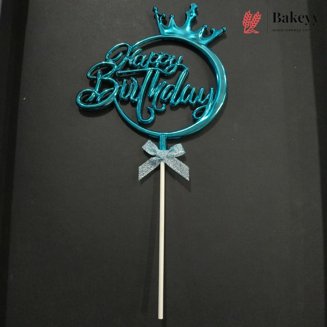 A metallic turquoise blue 'Happy Birthday' cake topper with a crown on a stick, displayed against a dark background. A Bakeyy logo is visible in the top right corner.