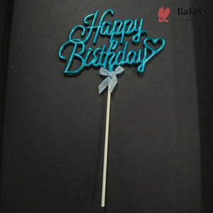 A turquoise blue glittery 'Happy Birthday' cake topper with a small silver bow on a white stick, set against a dark black background. A Bakeyy logo is visible in the top right corner.
