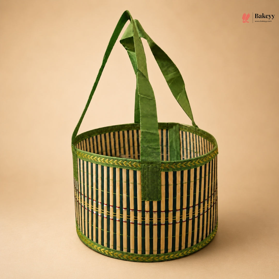 A round woven bamboo basket with green fabric handles, featuring vertical black and natural bamboo stripes with green braided trim, set against a solid beige background.