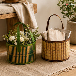 Two round woven bamboo baskets with handles, styled on a beige rug in a warm, rustic room. The left basket is green and filled with white flowers and greenery, while the right basket is natural tan and holds folded cream textiles and a magazine.