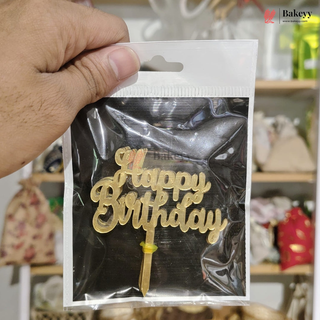 Gold 'Happy Birthday' cake topper in clear packaging held by a hand with a blurred background.