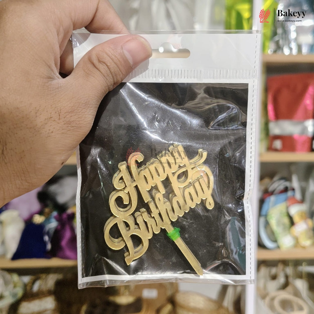 Gold 'Happy Birthday' cake topper in a clear plastic bag held by a hand.
