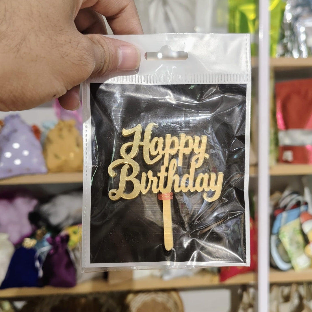 Gold 'Happy Birthday' cake topper in clear packaging held by a hand with a blurred background.