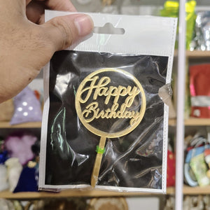 Gold 'Happy Birthday' cake topper in clear packaging held by a hand with a blurred background.