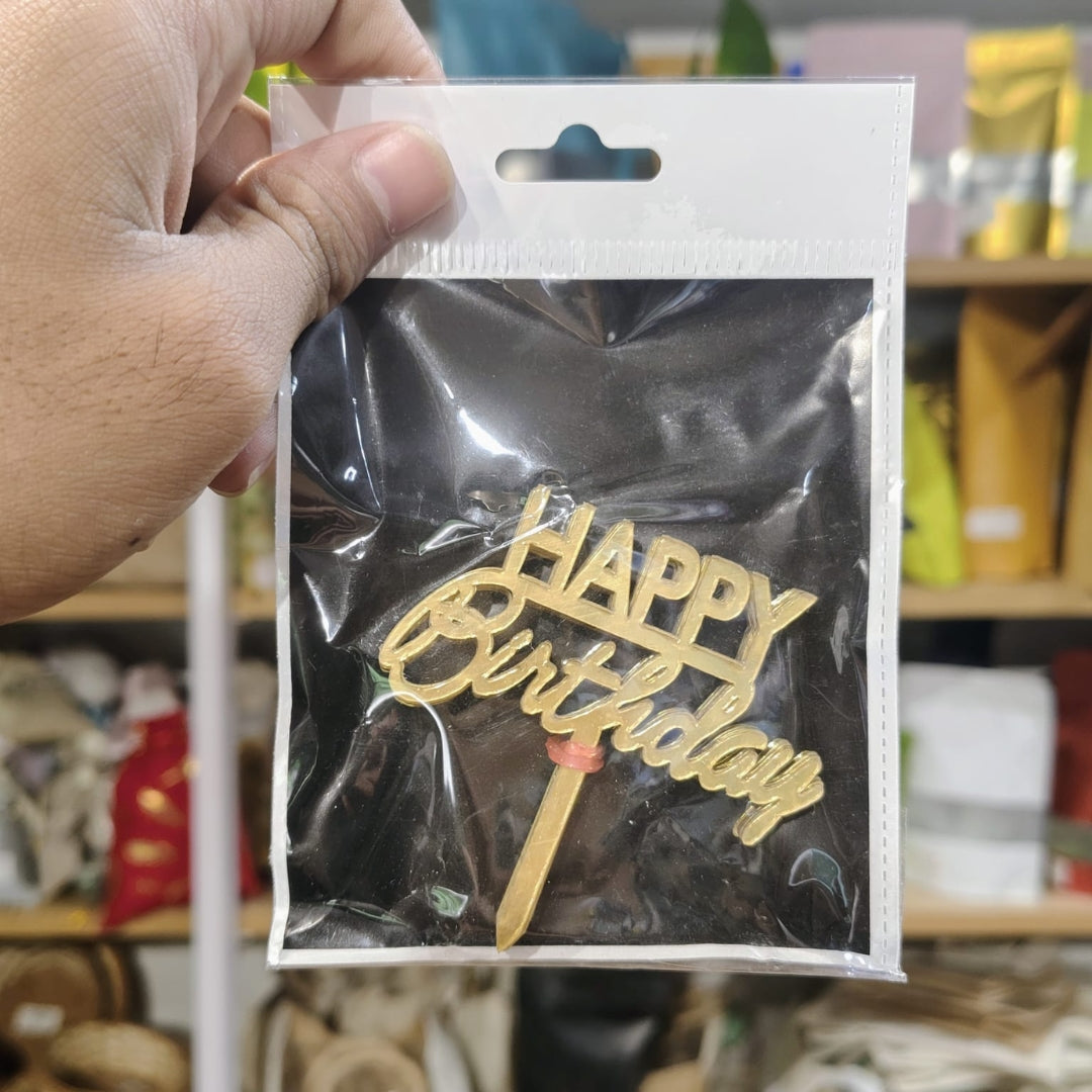 Gold 'Happy Birthday' cake topper in a clear package held by a hand with a blurred background.