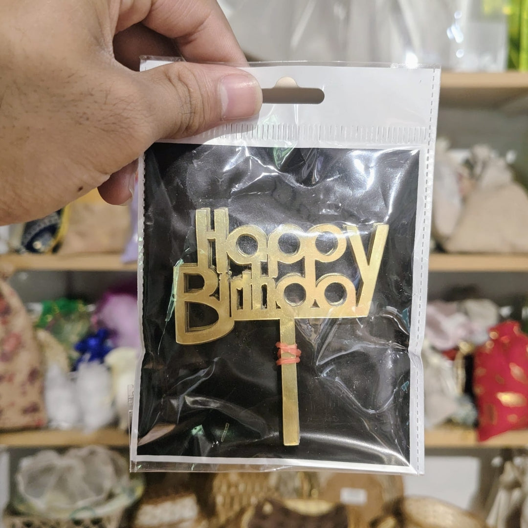Gold 'Happy Birthday' cake topper in a clear package held by a hand with a blurred background.