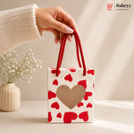 Hand holding a small gift bag with red hearts and a beige heart on a neutral background, featuring the Bakey brand.