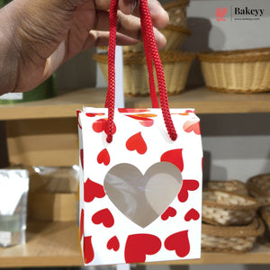 Hand holding a small gift bag with red heart patterns, with 'Bakeyy' branding in the corner.