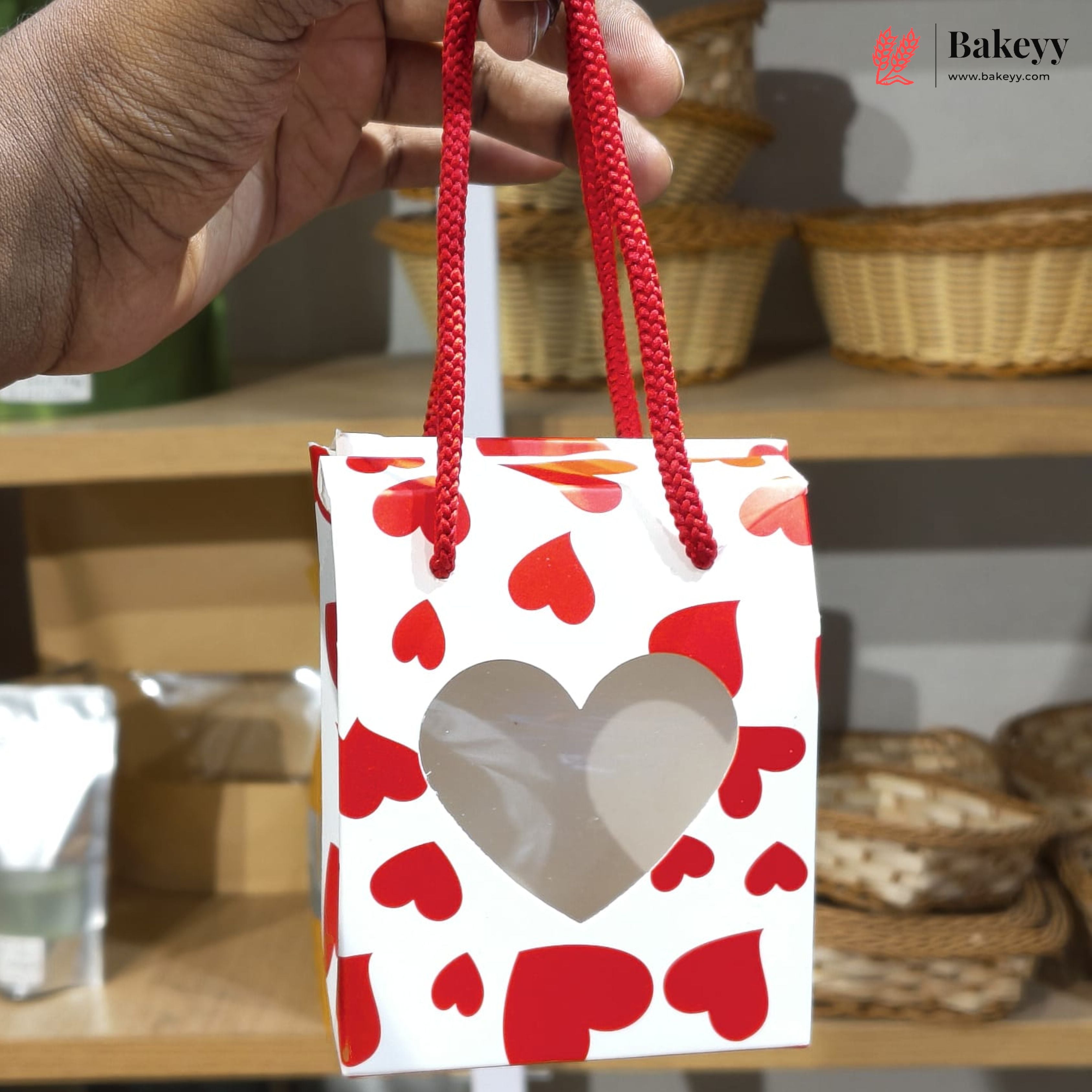 Hand holding a small gift bag with red heart patterns, with 'Bakeyy' branding in the corner.