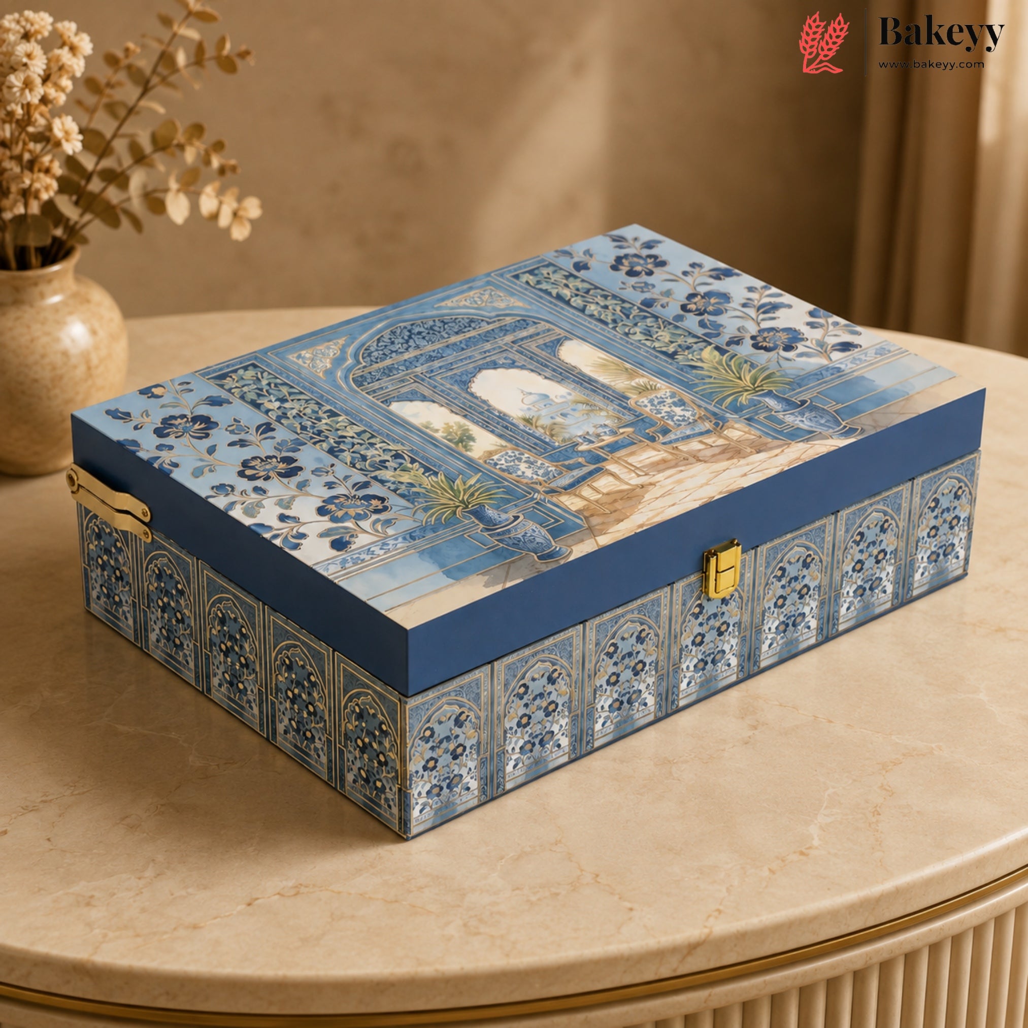 A rectangular decorative gift box with a blue and white architectural pattern on the lid and sides, featuring gold-tone latches, placed on a beige marble table in a warm indoor setting.