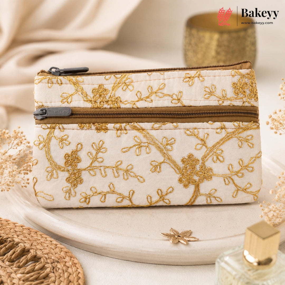 A cream-colored zippered pouch with gold floral embroidery, styled on a light beige ceramic plate with dried flowers and a perfume bottle in the background. The Bakeyy logo is visible in the top right corner.