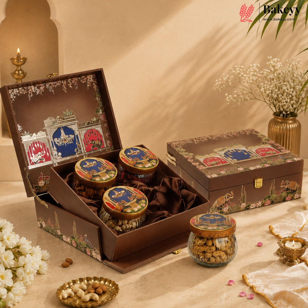 An open and closed decorative gift box containing three glass jars of nuts, arranged on a beige surface with flowers, candles, and a warm neutral background. The Bakey brand logo appears in the top right corner.