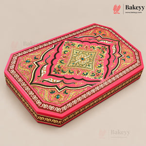 An ornate rectangular cake box with a vibrant pink lid decorated with intricate gold, green, and black traditional patterns, shown on a neutral beige background.
