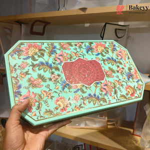 A hand holds a decorative octagonal mint green tin box with a colorful floral pattern and a red central medallion. The background shows a bakery shelf with bags and products.