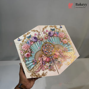 A hand holds a decorative hexagonal cardboard box with an intricate pastel illustration featuring fans, flowers, butterflies, and gold ornamental details. The background is a plain gray studio-like setting, and the Bakeyy logo appears in the top right corner.