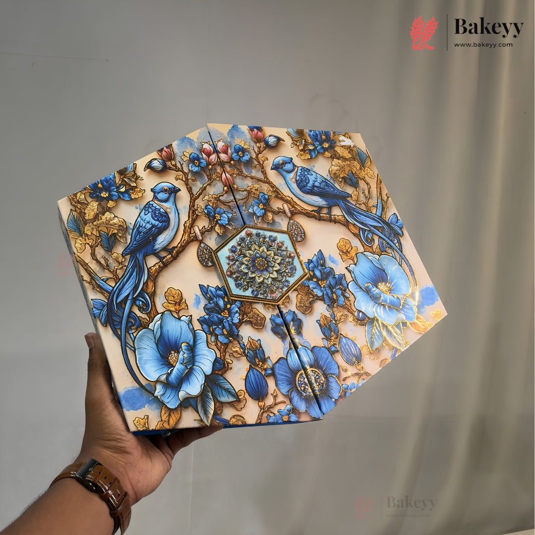 A hand holds a decorative, octagonal metal cake tin featuring an intricate illustration of blue birds, blue flowers, and gold-toned branches on a cream background. The Bakeyy brand logo is visible in the top right corner.