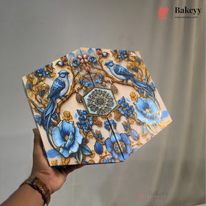 A hand holds a decorative, octagonal metal cake tin featuring an intricate illustration of blue birds, blue flowers, and gold-toned branches on a cream background. The Bakeyy brand logo is visible in the top right corner.