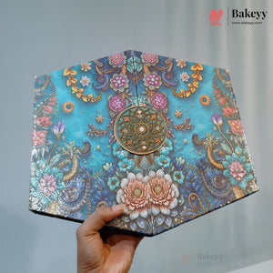 A hand holds a decorative, fan-shaped cake or pastry cover with an intricate turquoise floral and paisley pattern, gold accents, and a central medallion, set against a neutral gray background.