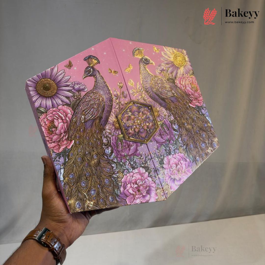 A hand holds a hexagonal pink decorative box featuring illustrated peacocks, flowers, and butterflies with gold foil accents. The background is a plain light gray fabric backdrop, and a Bakeyy watermark/logo appears in the top right corner.