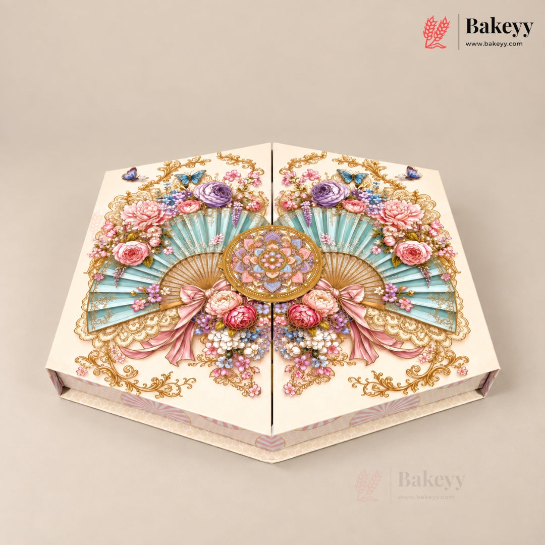 An ornate rectangular cake or pastry box with elaborate 3D floral and fan-like embroidery in pastel pink, lavender, turquoise, and gold on an ivory base, photographed on a warm beige background. The Bakeyy logo appears in the top-right corner.