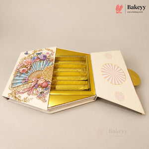An ornate open gift box for a baked good, featuring a cream-colored exterior decorated with a large pastel fan illustration, floral motifs, and gold detailing. Inside, several rectangular golden-yellow items are arranged in rows on a gold tray.