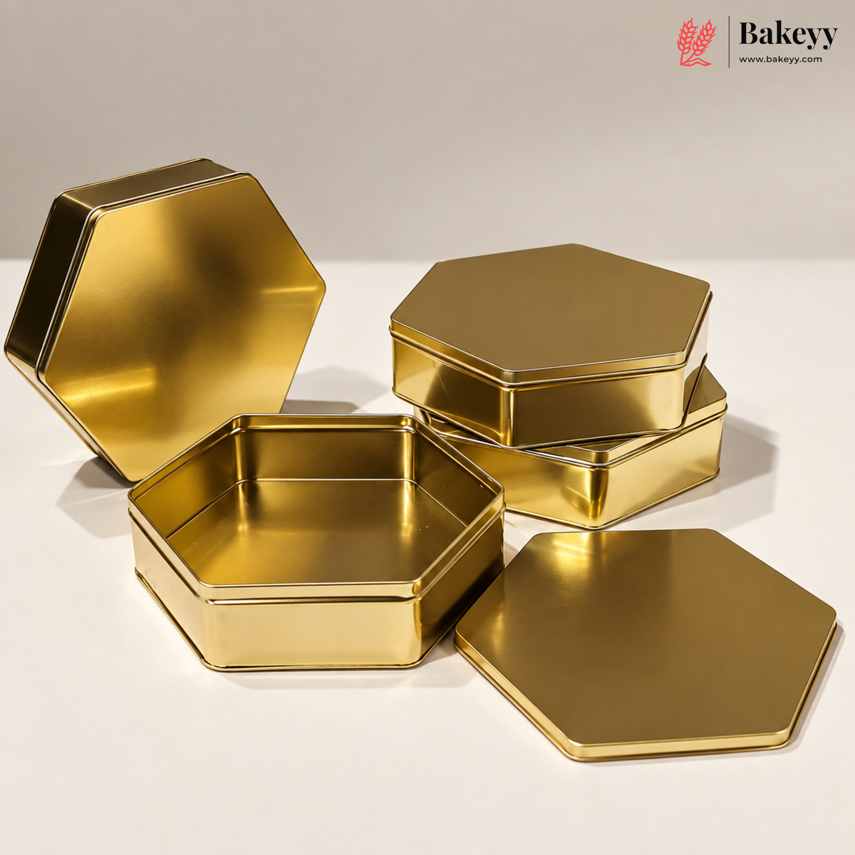 Five gold hexagonal metal tins and lids displayed on a clean white studio background, with the Bakeyy logo in the top right corner.