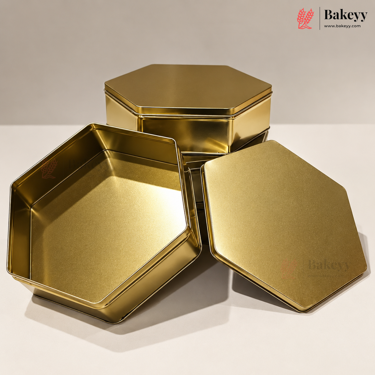 Three metallic gold hexagonal tin containers, including one open tin and two closed tins with lids, displayed on a light gray surface with a gray background. The Bakeyy logo is visible in the top right corner.