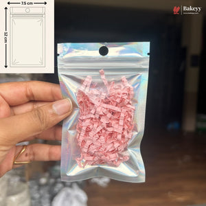 A hand holds a small holographic zip-top pouch filled with pink shredded paper against a dark indoor background. A white inset in the top left shows a simple packaging diagram labeled 7.