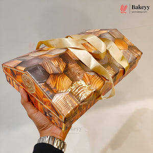 A hand holds a rectangular gift box wrapped in a metallic gold and brown geometric patterned paper with a shiny gold ribbon tied around it. The background is a plain light gray wall.