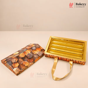 A rectangular chocolate gift box shown open, with a lid featuring a realistic chocolate chip pattern and a shiny gold interior tray. The box has a warm brown and orange patterned exterior and a gold ribbon handle, photographed on a clean beige studio background.