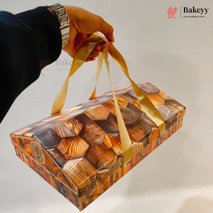 A hand wearing a silver watch holds a rectangular bakery gift box decorated with realistic wood-grain patterns in shades of brown and gold, complete with two gold ribbon handles, against a plain white background.