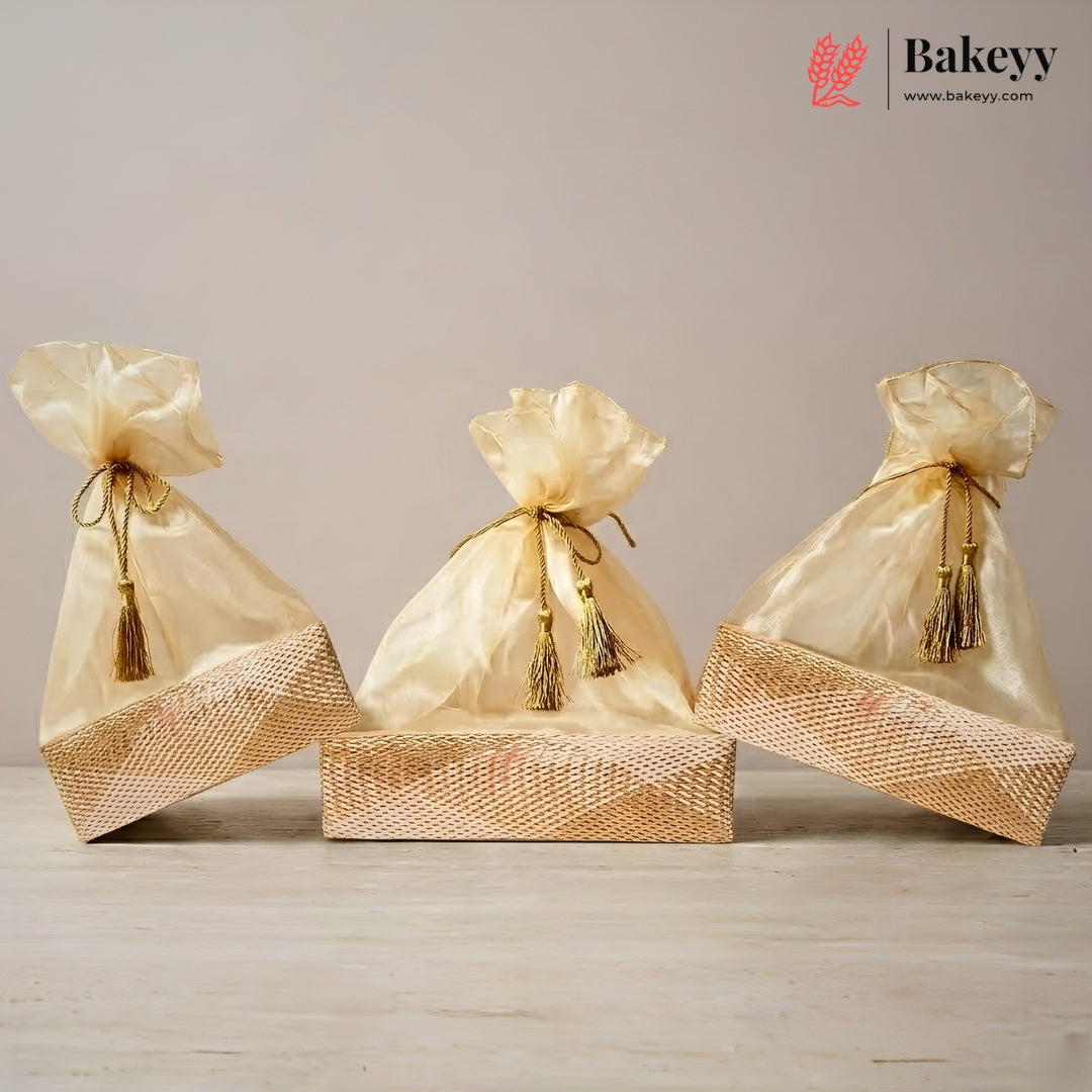 Three gold satin drawstring gift bags with tassel ties, arranged on top of a woven gold box against a neutral beige background. The Bakeyy logo is visible in the top right corner.