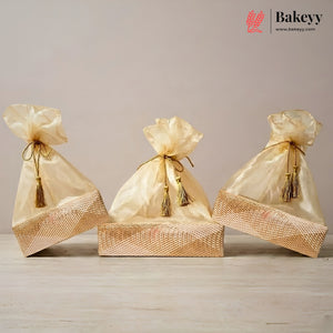 Three gold satin drawstring gift bags with tassel ties, arranged on top of a woven gold box against a neutral beige background. The Bakeyy logo is visible in the top right corner.
