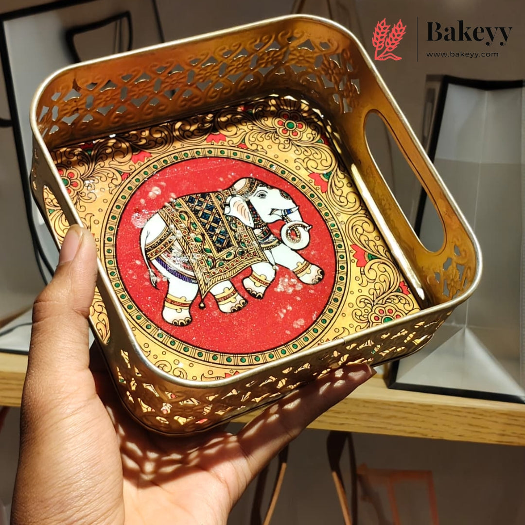 A hand holds a square decorative tray with cutout edges and built-in handles. The tray has a gold-toned ornate border and a central red panel featuring a painted elephant with white, black, and green details.