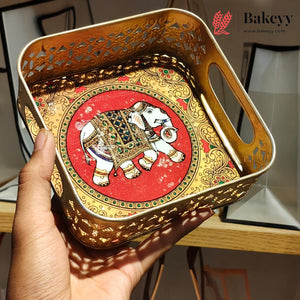 A hand holds a square decorative tray with cutout edges and built-in handles. The tray has a gold-toned ornate border and a central red panel featuring a painted elephant with white, black, and green details.