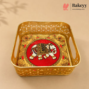 A square gold-colored metal tray with decorative cutouts and side handles, featuring an ornate red and gold elephant motif in the center. The Bakeyy brand logo is visible in the top right corner.