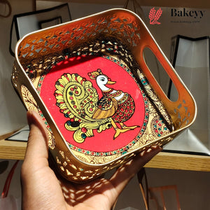 A hand holds a square decorative tray with cutout floral edges and built-in handles. The tray has a bright red inner panel featuring a colorful peacock illustration, with gold-toned metal trim and patterned detailing.